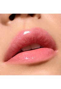 Prodotto labbra brillante con una sfumatura rosa chiaro, texture liscia e un leggero luccichio. Mostra labbra piene con un aspetto naturale e denti bianchi in vista.