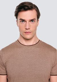 Brązowy, teksturowany t-shirt z okrągłym dekoltem i kontrastującym, ciemnobrązowym kołnierzem. Materiał wydaje się miękki i lekki.