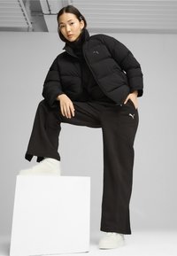 Sort pufferjakke med høj krave, sorte oversized bukser og hvide platform sneakers står på en hvid pedestal mod en grå baggrund.