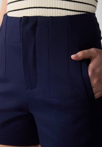 Kvinna som bär marinblå högmidjade shorts med vertikala sömdetaljer, handen placerad i höger ficka och en randig kräm- och svart topp.