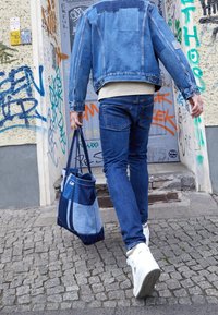 Džínová bunda a džíny v různých odstínech modré, doplněné vícebarevnou džínovou taškou, nošené s bílými teniskami na dlážděné stezce.