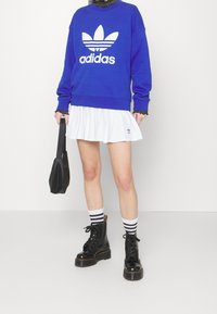 Blå sweatshirt med vit Adidas-logga, vit plisserad kjol, svarta lace-up stövlar med platå, och svart handväska. Randiga strumpor synliga.