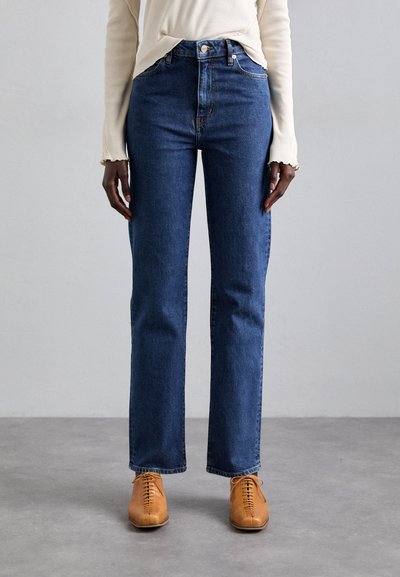 Persoon in hoge taille rechte spijkerbroek, beige lange mouwen top en tan leren veterschoenen staat op een grijze vloer.