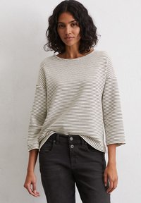 Long-sleeve, striped top in beige and black, featuring a wide neckline and relaxed fit. Paired with black jeans for a casual look.