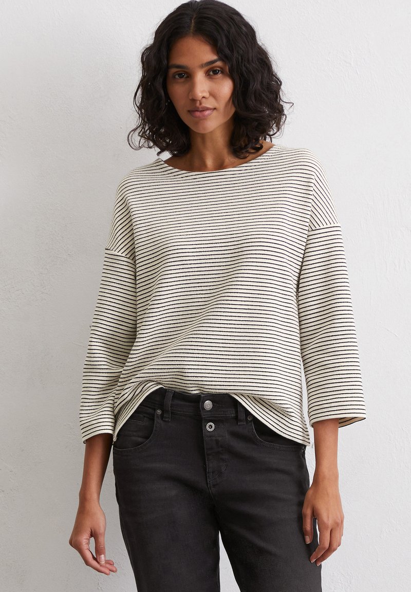Long-sleeve, striped top in beige and black, featuring a wide neckline and relaxed fit. Paired with black jeans for a casual look.