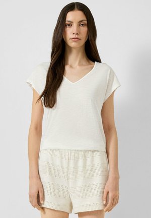 Femme aux longs cheveux foncés portant un haut blanc à manches courtes et col en V ainsi qu'un short en tricot crème texturé, debout devant un fond clair uni.