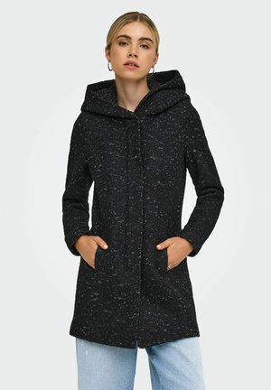 Femme portant un manteau à capuche noir moucheté, les mains dans les poches et un jean bleu clair, sur un fond clair uni.
