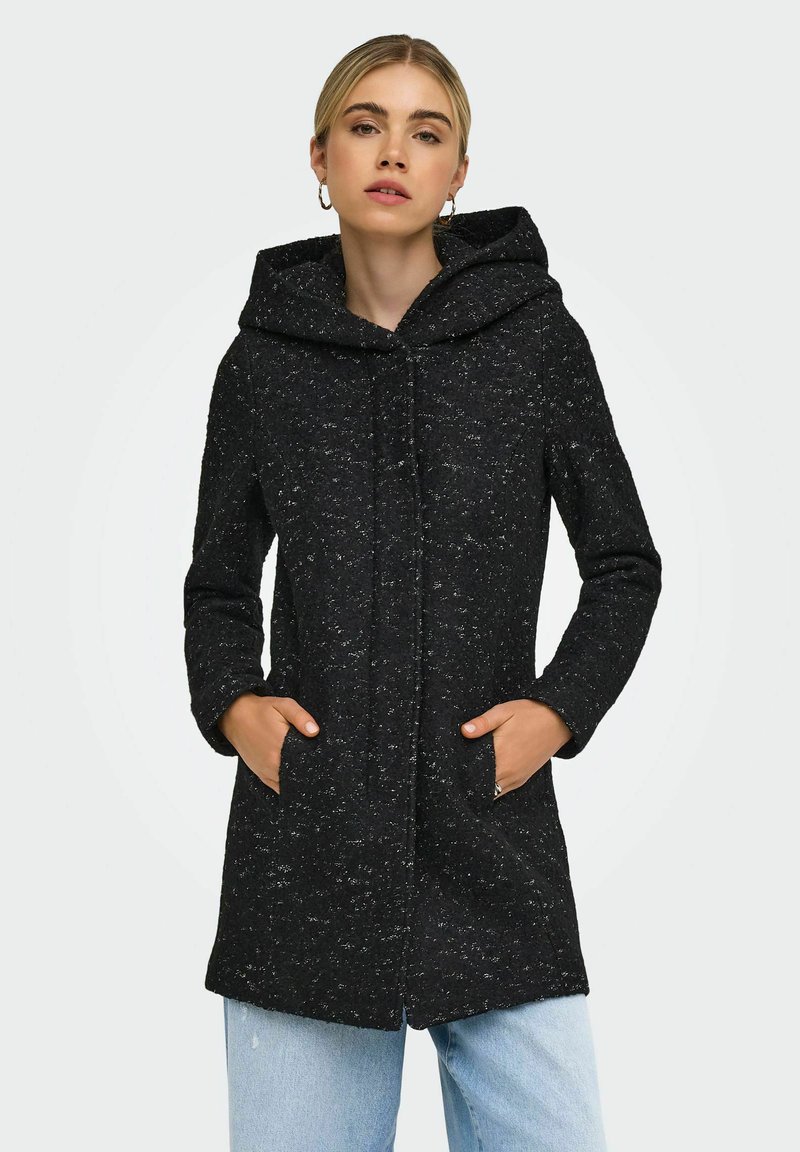 Femme portant un manteau à capuche noir moucheté, les mains dans les poches et un jean bleu clair, sur un fond clair uni.