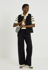 Chaleco negro de peluche sobre una camiseta tipo polo a rayas en crema y negro, combinado con pantalones negros de pierna ancha y zapatillas deportivas beige. Sosteniendo un bolso con estampado de cebra.