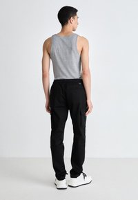 Graues ärmelloses Tanktop kombiniert mit schwarzen Cargo-Hosen mit mehreren Taschen. Modell trägt weiße Sneaker. Rückansicht, minimalistischer Hintergrund.