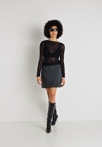 Čierny čipkovaný top s dlhými rukávmi skombinovaný so šedou mini sukňou a čiernymi vysokými podpätkami po kolená. Modelka s tmavými slnečnými okuliarmi stojí na neutrálne pozadí.