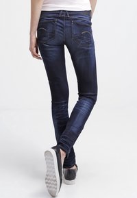 Jeans ajustados en denim de lavado oscuro con una textura suave, presentando un desvanecimiento sutil, bolsillos traseros y detalles en costura dorada. Combinados con zapatos de lona negros sin cordones.