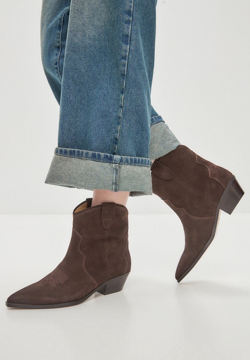 Persona che indossa jeans blu risvoltati e stivaletti in suede marroni con tacco basso su un pavimento chiaro.