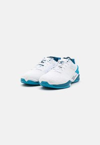 Des chaussures de sport blanches avec une tige texturée, des accents bleus au niveau du talon et de la semelle, et une semelle intermédiaire rembourrée. Elles présentent un laçage traditionnel.