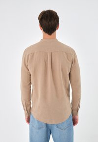 Cămașă lungă bej deschis, cu guler, detalii cu pliu pe spate și manșete cu nasturi. Asortată cu blugi din denim albastri.