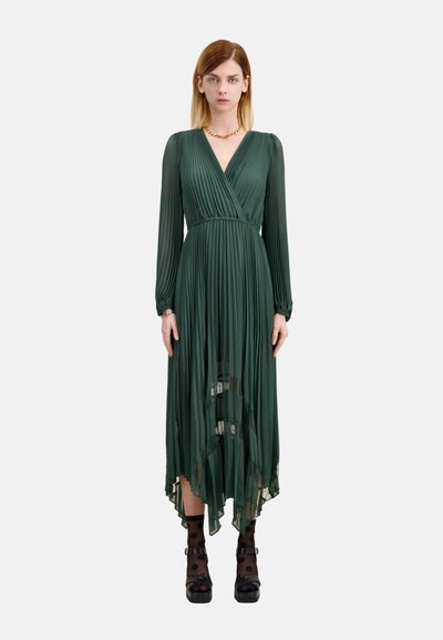 Zielona sukienka z plisowaniem, z dekoltem w serek, długimi rękawami i zwiewnym dołem. Akcentowana przez przezroczysty materiał u dołu i zestawiona z czarnymi butami na platformie.