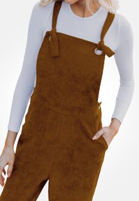 Pantalones de tirantes de pana marrón con tirantes anudados, bolsillos laterales y cintura ajustada. Debajo, se ve una camiseta de manga larga ligera.