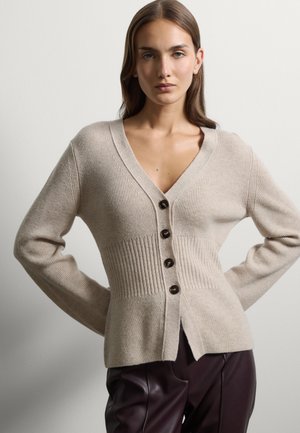 Junge Frau trägt eine beige geknöpfte, gerippte Strickjacke und dunkelbraune Lederhose, steht mit den Händen in der Hüfte vor einem einfarbigen Hintergrund.