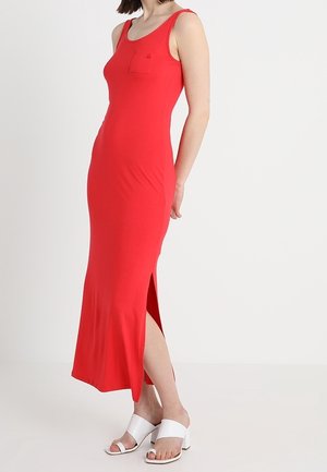 Nainen, joka pukeutunut hihattomaan kirkkaan punaiseen maksimekkoon, jossa on sivuhalkio, yhdistettynä valkoisiin avonaisiin sandaaleihin, seisoo yksinkertaisen taustan edessä.