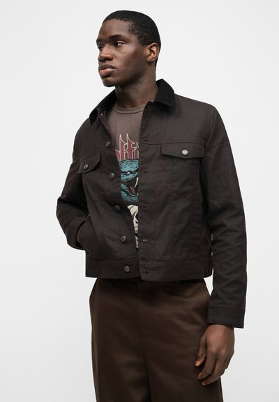 Άνδρας με σκούρο καφέ σακάκι με κουμπιά, φορώντας ένα γραφικό μπλουζάκι με μπλε σχέδιο φιδιού, στέκεται με το ένα χέρι στην τσέπη μπροστά σε λευκό φόντο.