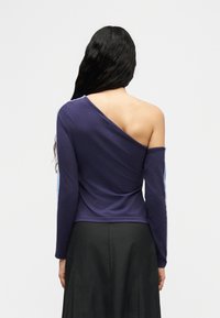 Γυναίκα με μακριά σκούρα μαλλιά φοράει ασύμμετρο μπλε σκούρο τοπ που αφήνει ακάλυπτο τον δεξί της ώμο και μαύρη φούστα, στραμμένη πλάτη σε απλό φόντο.