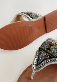 I sandali adornati presentano una fascia argentata con perline multicolori, accentuata da pietre trasparenti e metalliche. La suola è marrone con una finitura testurizzata.