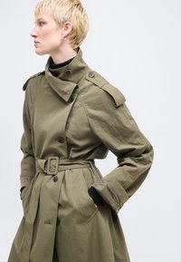 Άτομο με κοντά ξανθά μαλλιά που φοράει χακί πράσινο παλτό με ζώνη και ψηλό γιακά, στέκεται με τα χέρια στις τσέπες, κοιτάζοντας προς τα αριστερά.