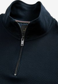 Tmavomodrý sveter s textúrovanou tkaninou, vysokým golierom a polovičným zipsom. Má strieborný zips a vzorovanú podšívku vnútorného goliera.