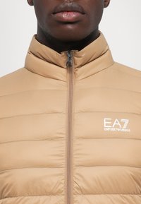 Bež puffer jakna s visokim ovratnikom, zatvaranjem na zatvarač, horizontalnim šavovima i EA7 Emporio Armani logotipom u bijeloj boji na lijevoj strani prsnog dijela.