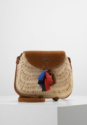 Bolso bandolera de ratán tejido con solapa de ante marrón y decoraciones de borlas coloridas en rojo, azul y negro sobre un pedestal blanco.