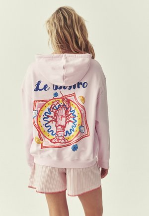 Kvinde med blondt hår iført en lyserød hættetrøje med en farverig hummer og teksten "Le bistro" på ryggen, kombineret med stribede shorts.