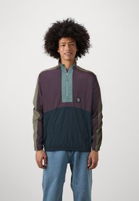 Pullover jacket in purple, teal, and dark navy. Features a half-zipper, elastic cuffs, and a logo patch on the front. Lightweight fabric.