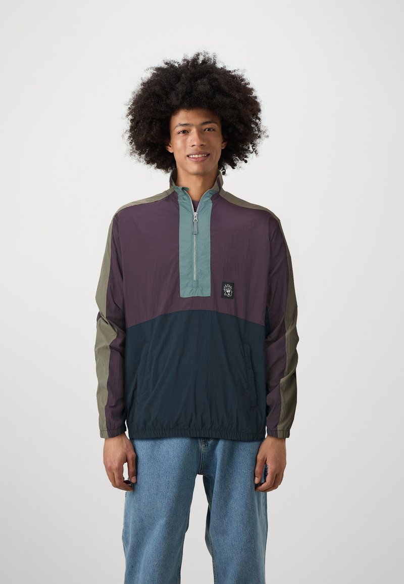 Pullover jacket in purple, teal, and dark navy. Features a half-zipper, elastic cuffs, and a logo patch on the front. Lightweight fabric.
