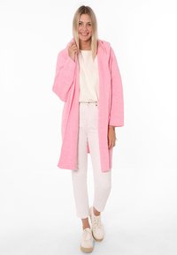 Rosa Kapuzenjacke mit langen Ärmeln, strukturiertem Stoff, offener Front, kombiniert mit hellen Hosen und weißen Sneakers.