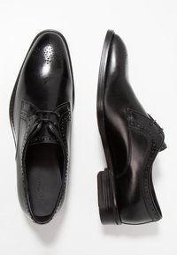 Chaussures habillées en cuir noir avec une finition lisse, des détails perforés et un petit talon empilé. Elles présentent un design à lacets et un bout arrondi.