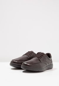 Een paar donkerbruine leren casual veterschoenen met gevoerde kraag en getextureerde rubberen zolen op een witte ondergrond.