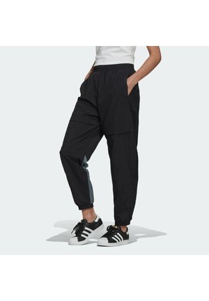 Personne portant un pantalon de jogging noir avec des poignets élastiques et des baskets Adidas noir et blanc, debout les mains dans les poches.