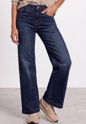 Personne portant un jean bleu foncé à jambes larges et des baskets beiges, debout avec une main dans la poche sur un fond neutre.