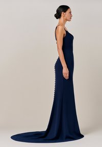 Temno modra večerna obleka z oprijetim zgornjim delom, tankimi naramnicami in tekočim vlečkom. Ponaša se z gumbi ob strani. Gladek tekstura.