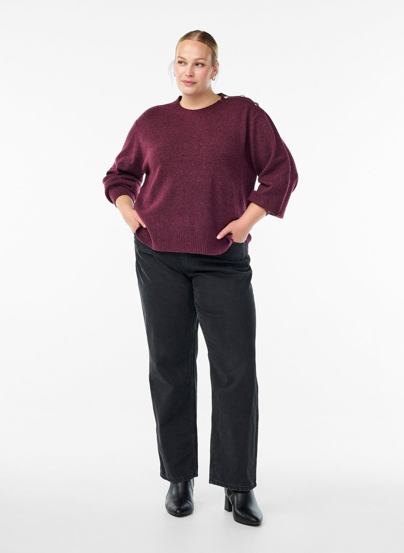 Kvinde med blondt hår i en burgunderrød sweater, sorte vidde jeans og sorte ankelstøvler, står med hænderne i lommerne på en hvid baggrund.