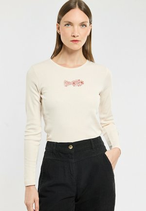 Lange mouw crème top met een ronde halslijn, voorzien van een rood vis- en bloemontwerp op de borst, gecombineerd met hooggerande zwarte corduroy broek.