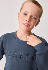Blaues Baumwoll-Sweatshirt mit geripptem Ausschnitt und Bündchen. Auf der Vorderseite ist ein erhabener Schriftzug "BAKER by TED BAKER" in dunklerem Blau zu sehen. Glatte Textur.
