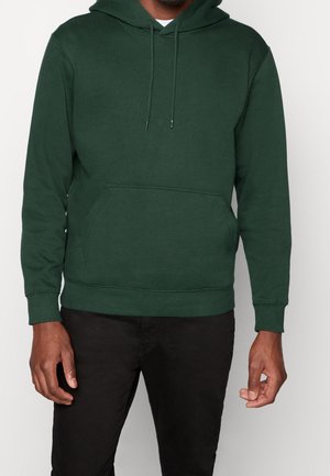 Homme portant un pull à capuche vert foncé avec poche avant, et un pantalon noir, debout devant un fond clair uni.