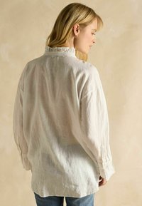 Woman with blonde hair wearing a loose white long-sleeve blouse with ruffled collar and blue jeans, facing away against a beige background.
