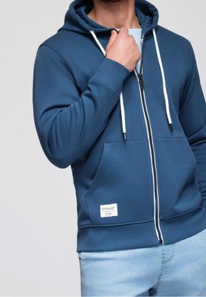 Mann trägt einen blauen Hoodie mit Reißverschluss und weißen Kordeln sowie hellblaue Jeans, hält den Hoodie am Hals.