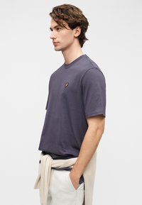 Μωβ βαμβακερό T-shirt με κοντά μανίκια, στρογγυλή λαιμόκοψη και μικρό κίτρινο κεντημένο λογότυπο στο στήθος, φορεμένο με ανοιχτόχρωμο παντελόνι.