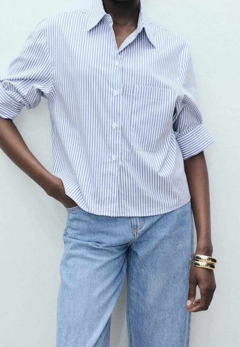 Personne portant une chemise boutonnée à rayures blanches et bleues avec les manches retroussées, un jean bleu clair et des bracelets en or au poignet droit.