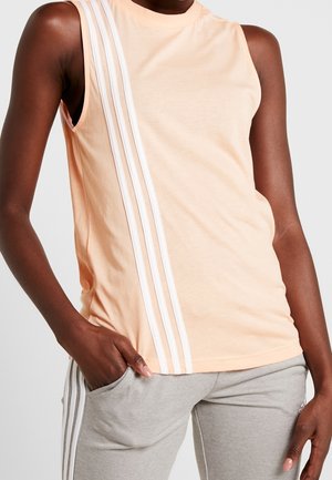 Bezruký broskvový top se třemi bílými svislými pruhy na levé straně, nošený s světlými šedými kalhotami se stejnými bočními pruhy.