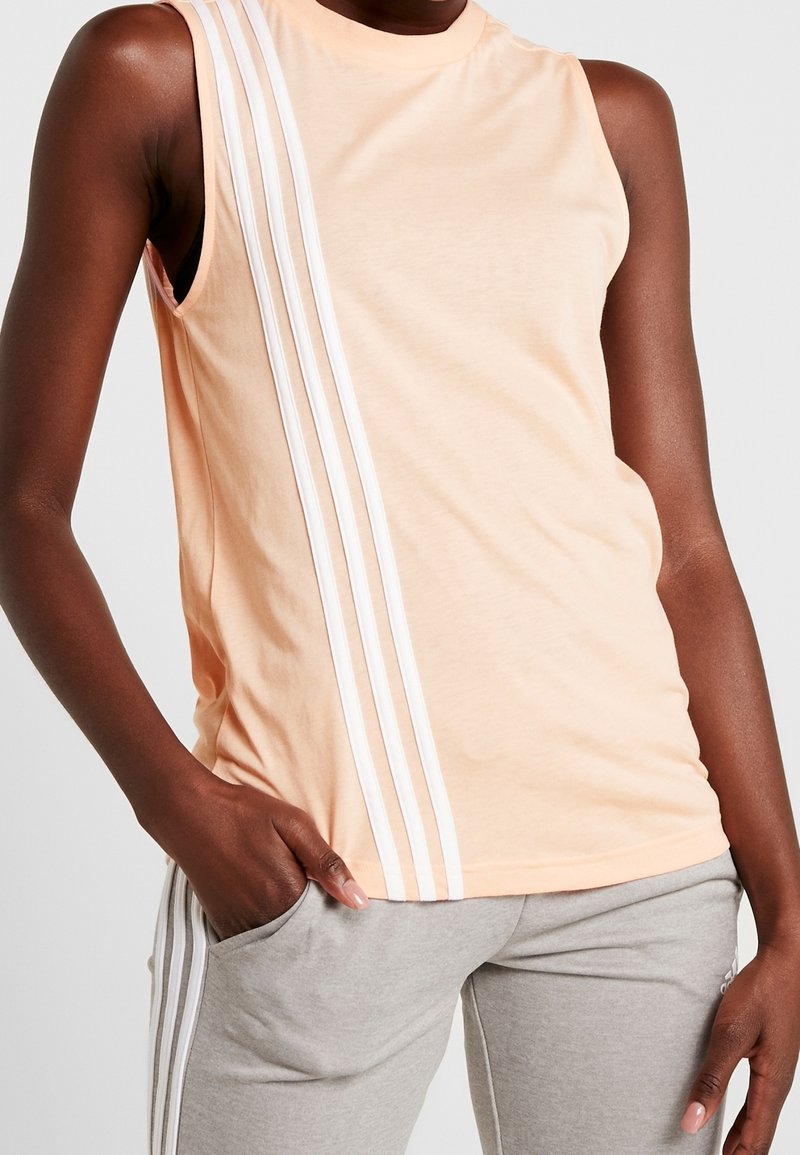 Haut sans manches couleur pêche avec trois rayures blanches verticales sur le côté gauche, porté avec un pantalon gris clair avec des rayures latérales assorties.