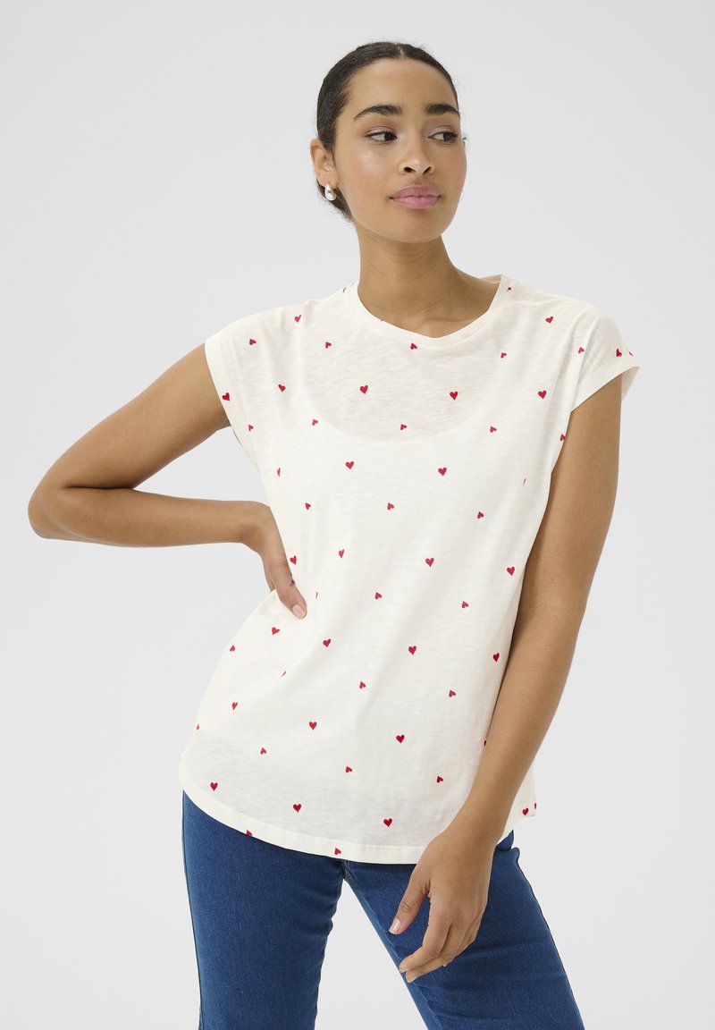 T-shirt crème à manches courtes avec un motif de cœurs rouges dispersés, col rond et coupe décontractée, associé à un jeans en denim bleu.